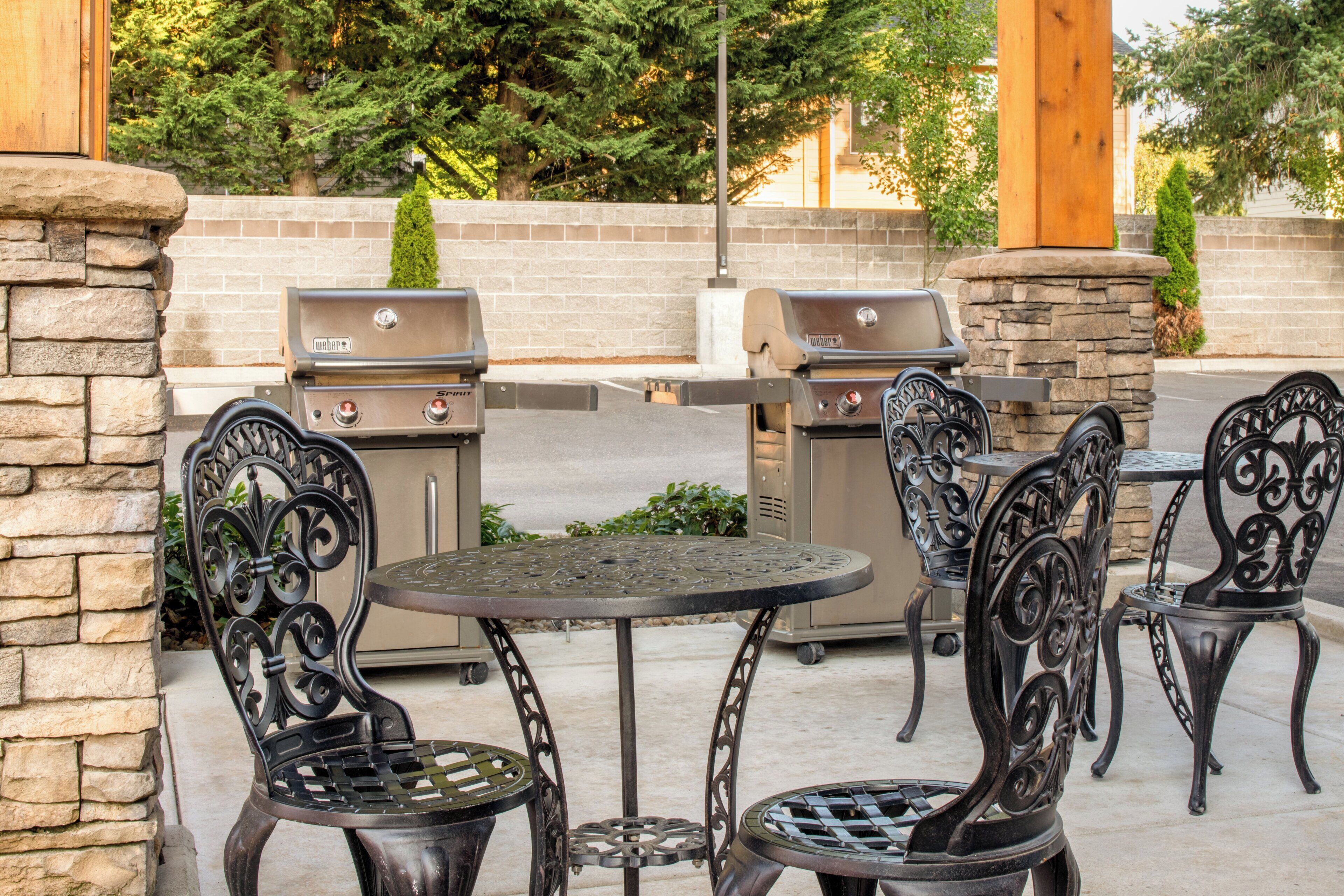 An outdoor patio with two Weber grills, stone pillars, and wrought iron chairs.