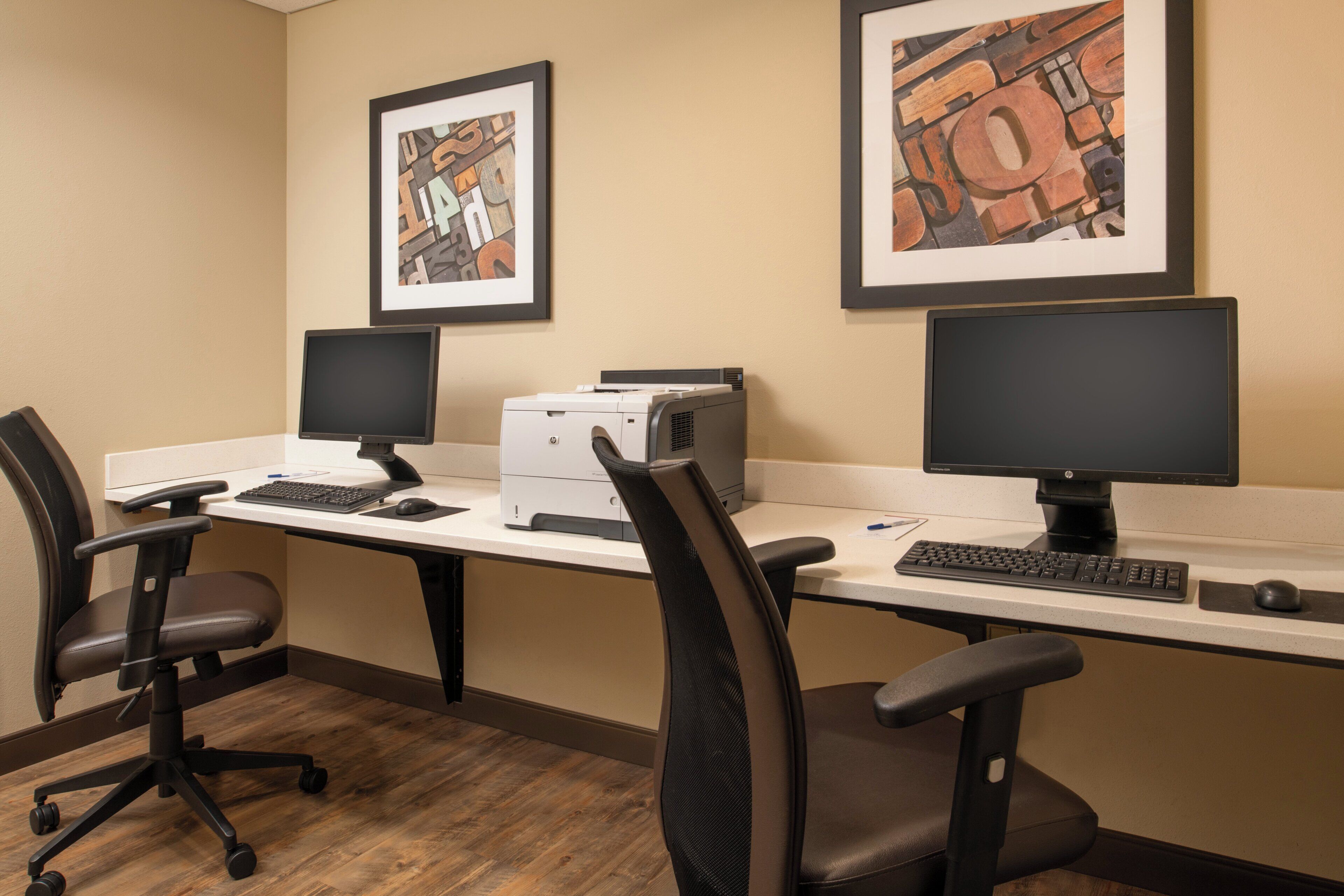 A workspace with two desks, each with a computer monitor, keyboard, and chair. There are framed artworks on the wall above the desks.
