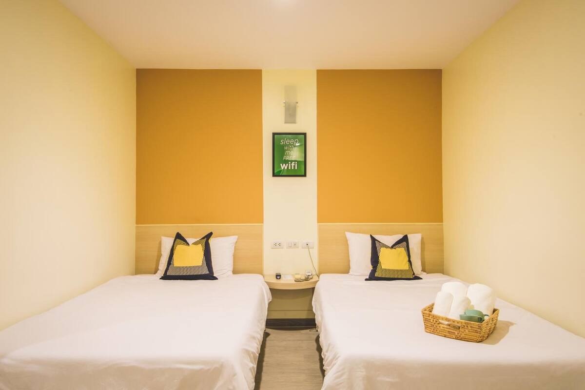Quarto de hotel com duas camas, uma mesinha e um cesto de toalhas.