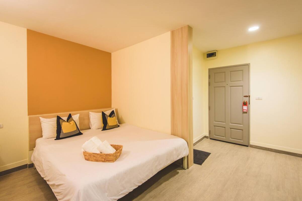 Quarto de hotel com uma cama grande, cabeceira de madeira, porta com alarme de incêndio e piso em tons claros.