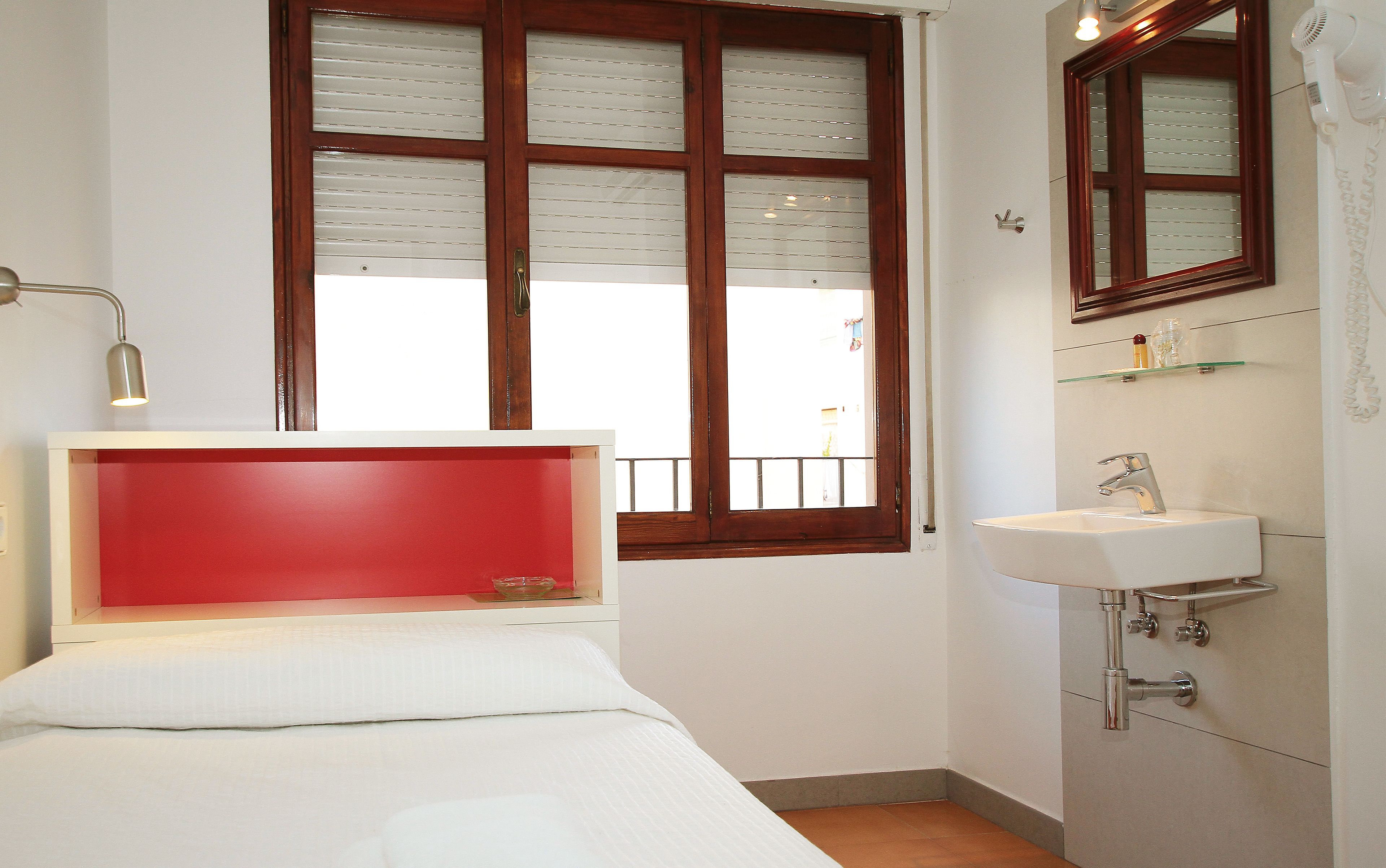 Un dormitorio con cama, una lámpara de noche, una ventana con persianas, un baño con lavabo y espejo, y una pared de acento roja.