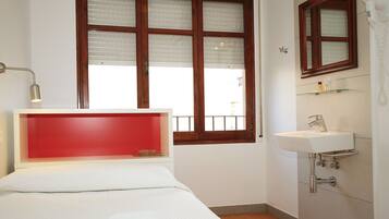Un dormitorio con cama, una lámpara de noche, una ventana con persianas, un baño con lavabo y espejo, y una pared de acento roja.