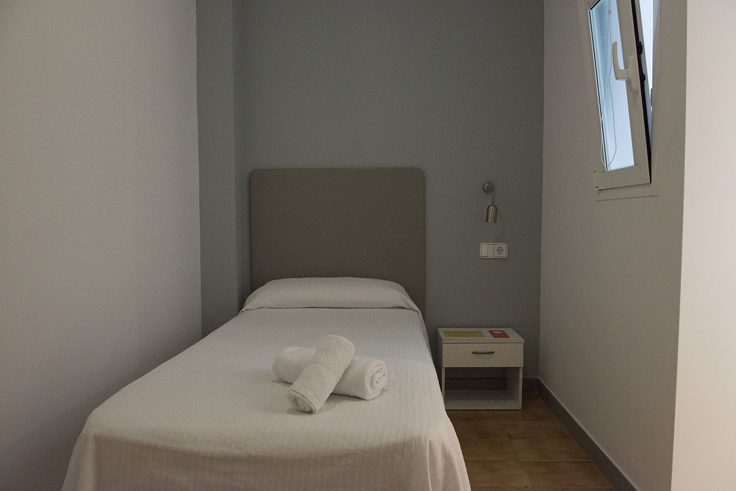 A small, single-bed room with a plain headboard, a bedside table with a lamp, and a window with a view.