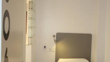 Una habitación pequeña y bien iluminada con una cama individual, una mesita de noche, un aire acondicionado instalado en la pared y una puerta con un número.