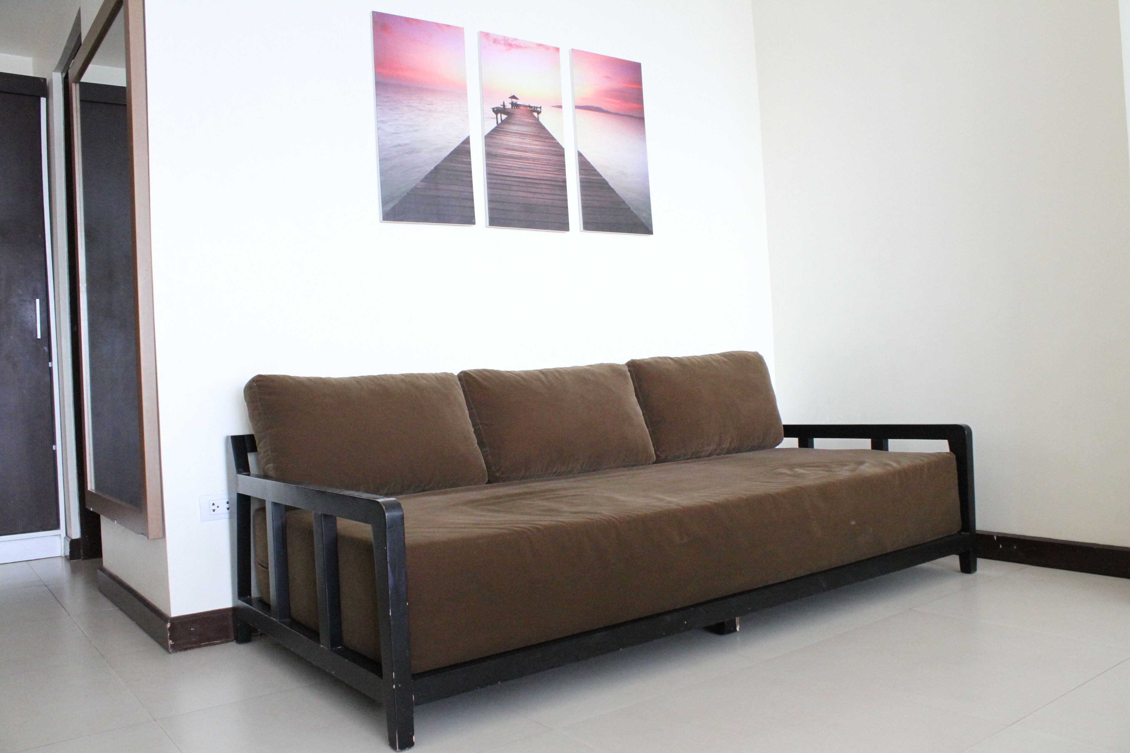 A brown sofa with a wooden frame in a room with a white wall and a wooden floor.