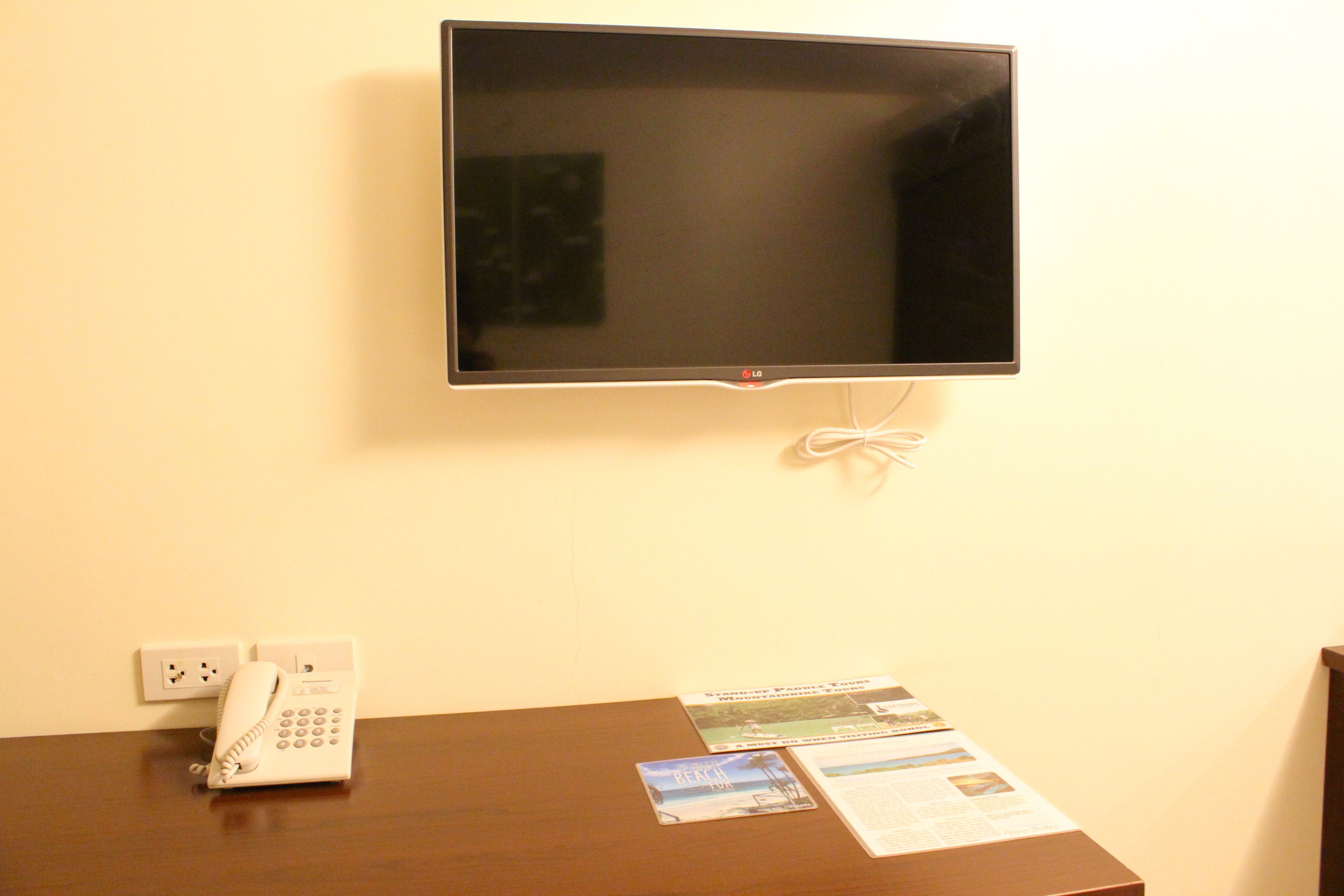 A hotel room with a flat-screen TV mounted on the wall, a telephone on the table, and some printed materials.