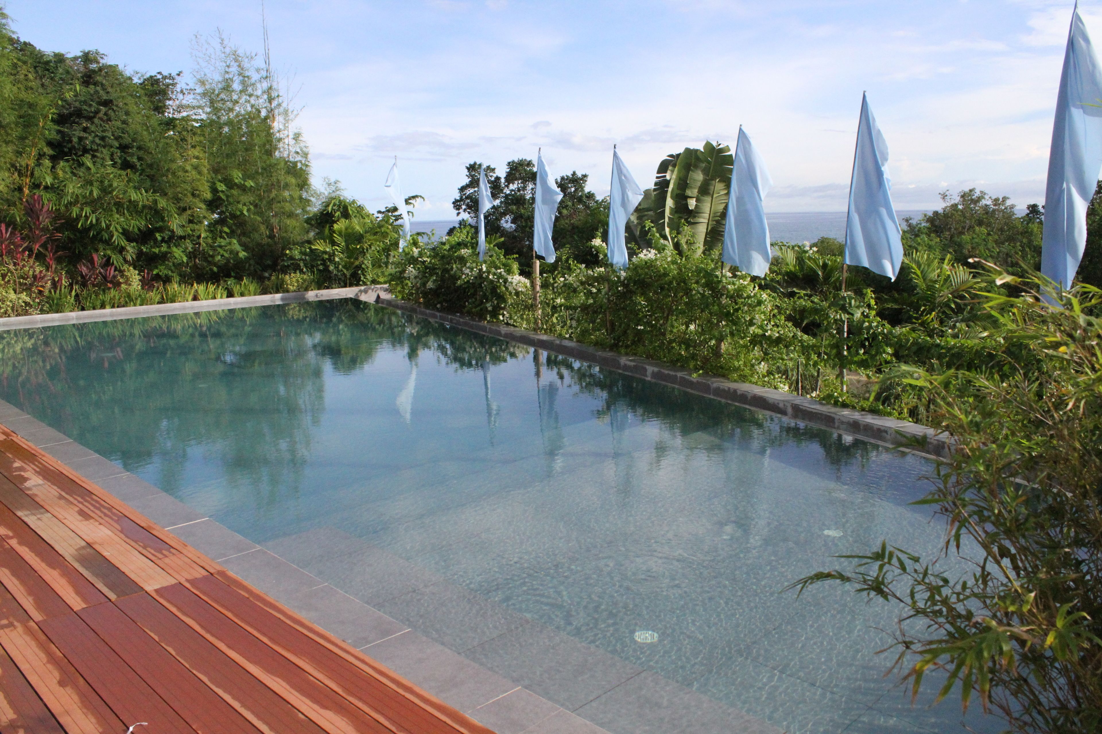 A rectangular infinity pool with a wooden deck on one side, surrounded by lush greenery and flags.