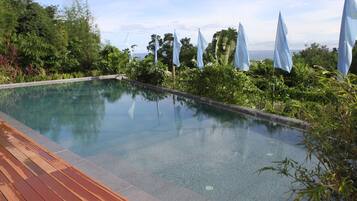 A rectangular infinity pool with a wooden deck on one side, surrounded by lush greenery and flags.