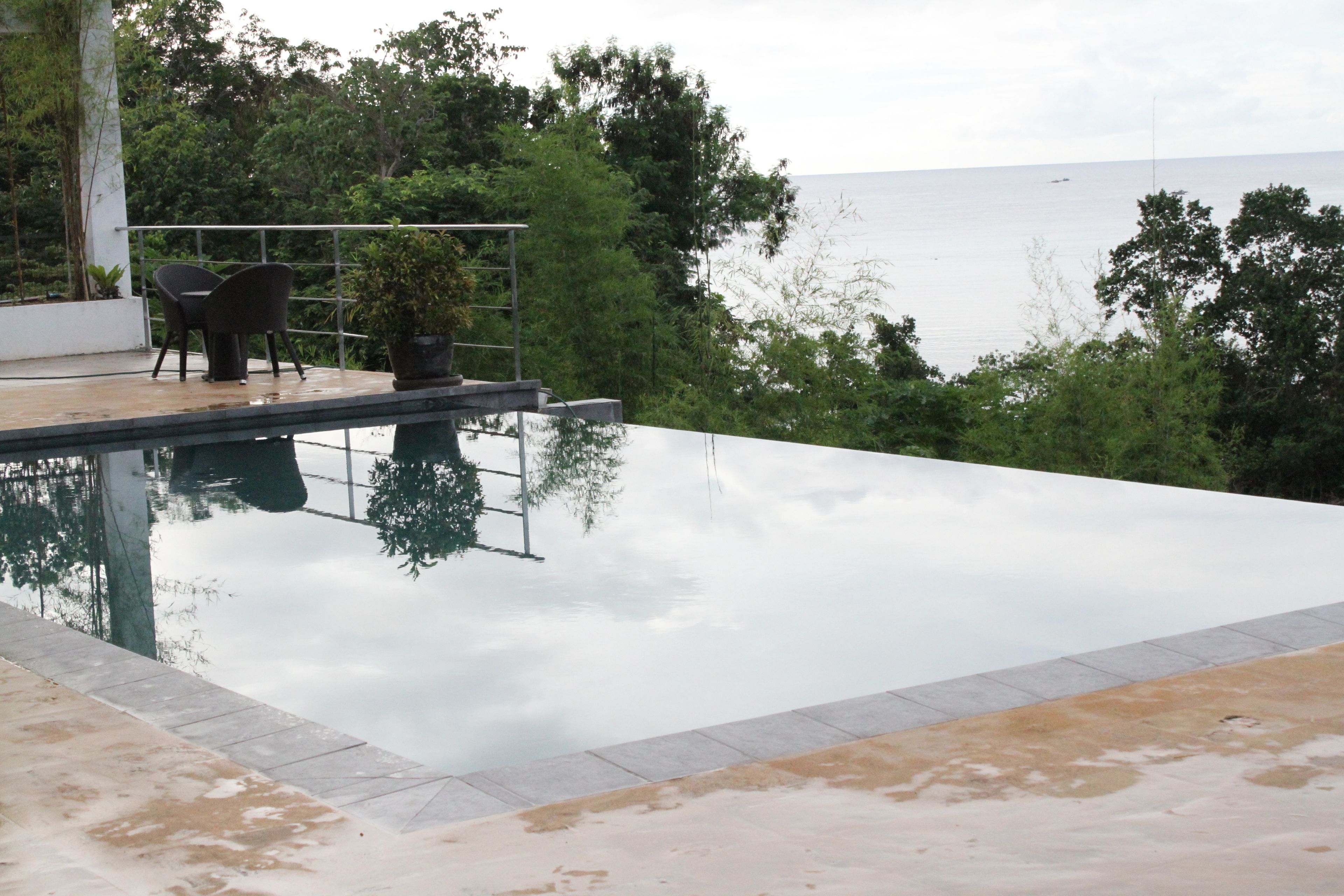 An infinity pool with a view of the sea, surrounded by greenery and a seating area with chairs.