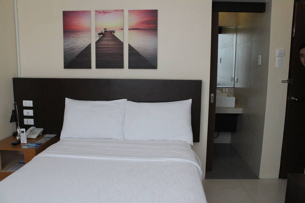 Ein Hotelzimmer mit einem großen Bett, einem Nachttisch mit Telefon und Zeitschriften sowie einer Wand mit drei Sonnenuntergangsbildern.