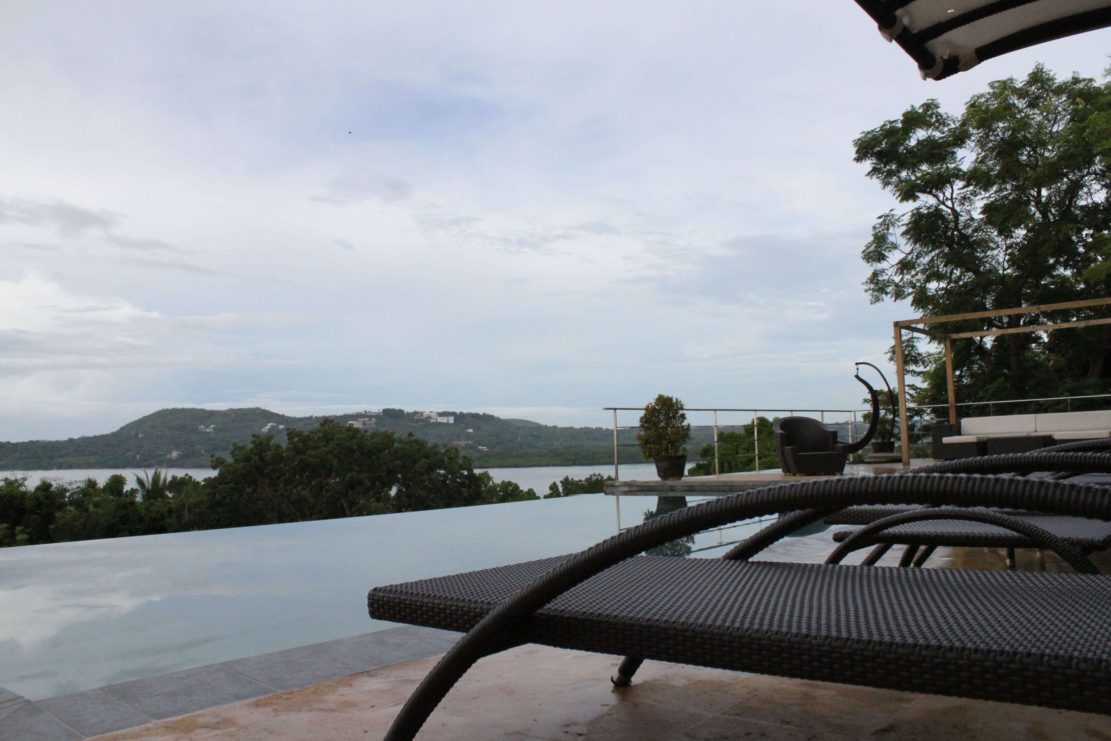 An infinity pool with a view of a lake and hills. There are wicker chairs and a potted plant.