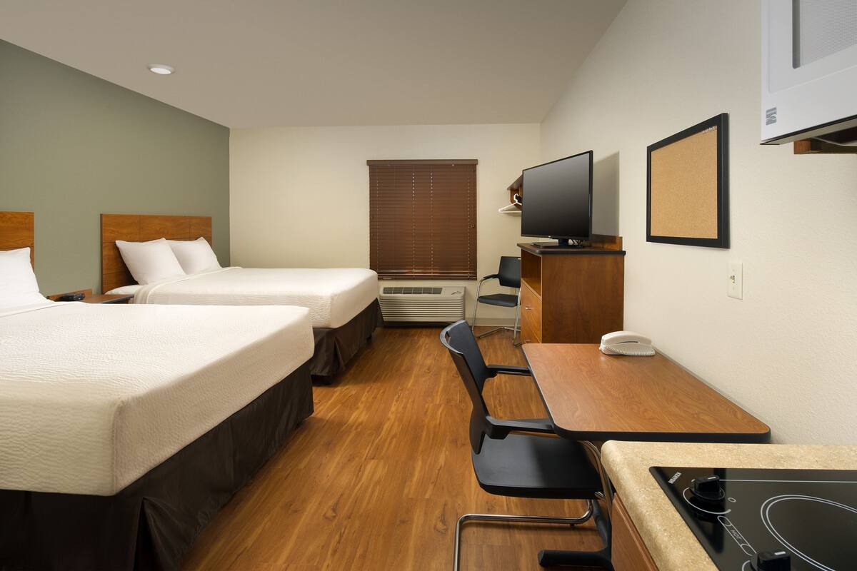 A hotel room with two beds, a desk, a chair, a television, and a corkboard on the wall.