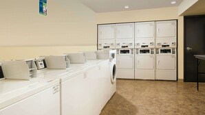 Un cuarto de lavandería con varias lavadoras y secadoras, piso de baldosas y una puerta a la derecha.