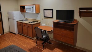 Cocina compacta con refrigerador, microondas, pileta y armarios de madera. Cuenta con una pequeña mesa de comedor con una silla y un televisor sobre un atril de madera.