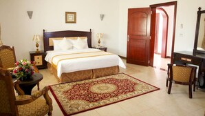 A hotel room with a large bed, two bedside tables with lamps, a wooden chair, a desk with a mirror, and a red patterned rug. - Green Mubazzarah Chalets (Al Ain)