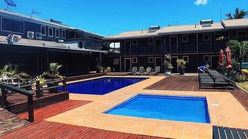 Uma área de piscina com deck de madeira, duas piscinas e um edifício com várias varandas.