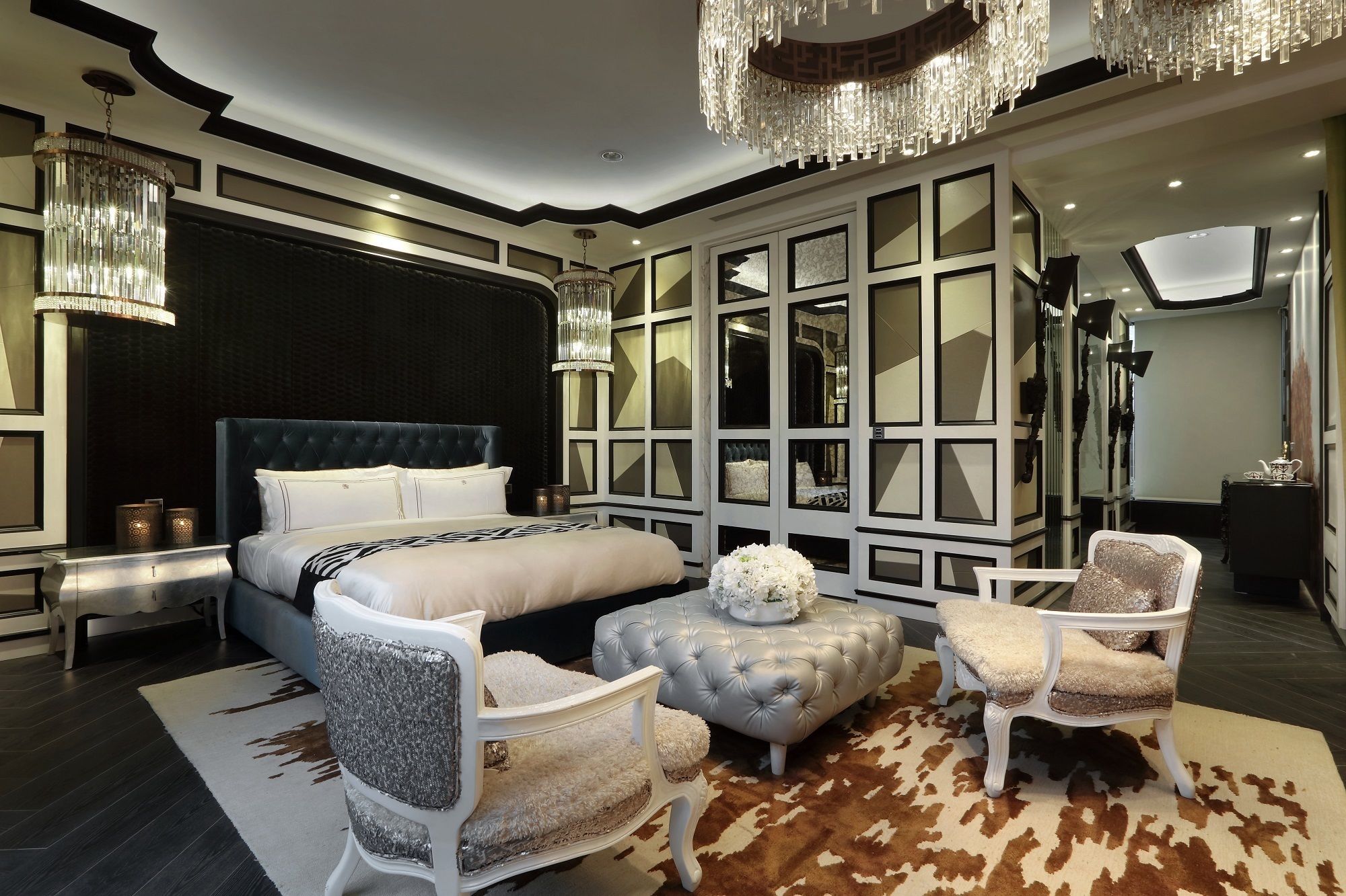 A modern bedroom with a large bed, two armchairs, a tufted ottoman, and a chandelier.