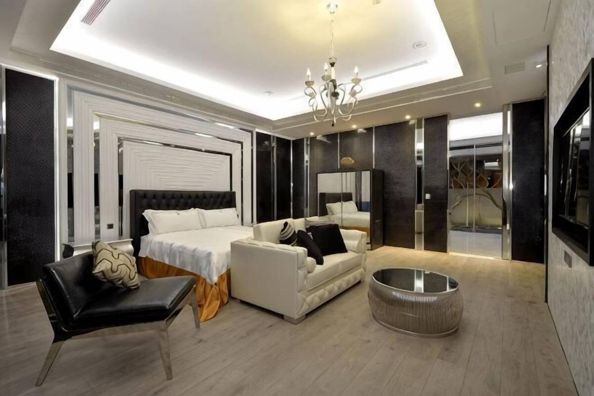 Un dormitorio moderno con una cama grande, un sofá, una silla, un reposapiés redondo y un televisor.