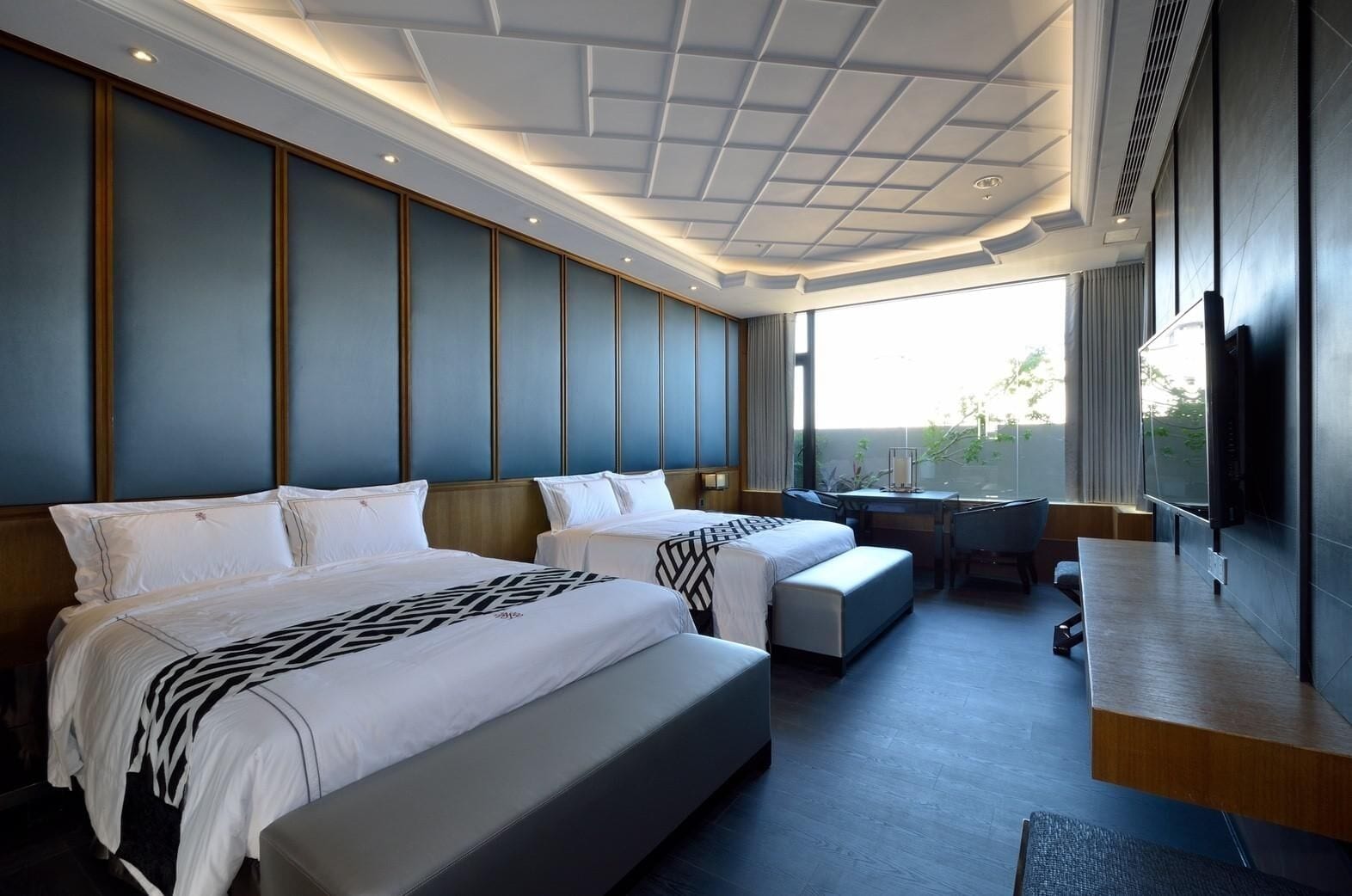 A hotel room with two beds, a desk, and a large window.