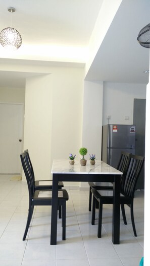 Un comedor con una mesa blanca y sillas negras, un colgante de luz y un refrigerador al fondo.