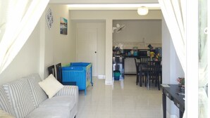 A living room with a sofa, a small table, a baby playpen, and a kitchen area in the background. - H&A Guest House (Malacca City)