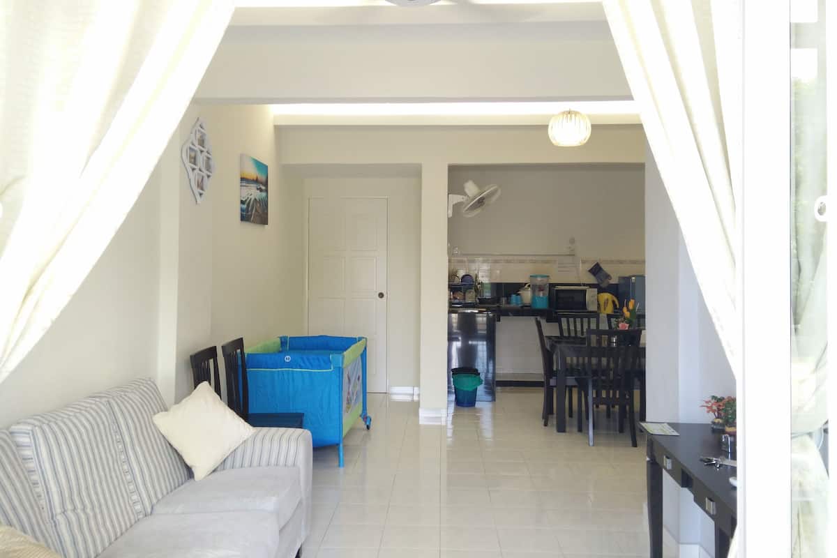 Uma sala de estar com sofá, uma mesa pequena, um cercadinho infantil e uma cozinha ao fundo.