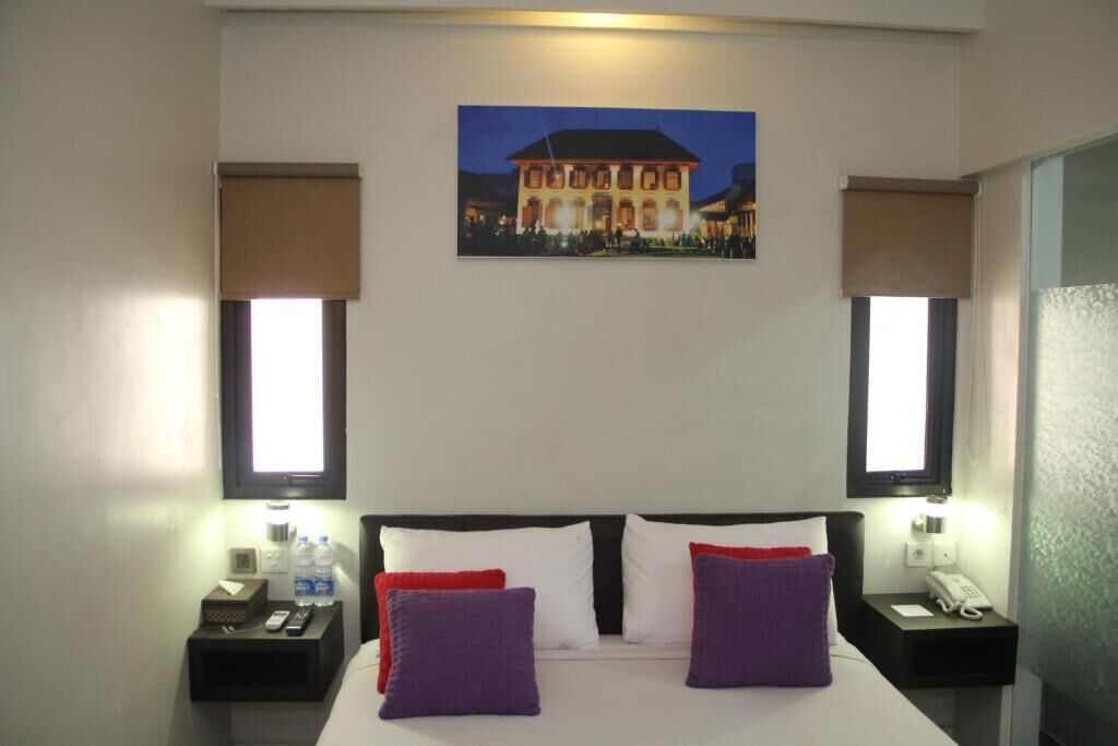 Habitación de hotel con una cama grande, dos mesitas de noche, un cuadro de un edificio y dos ventanas con persianas.