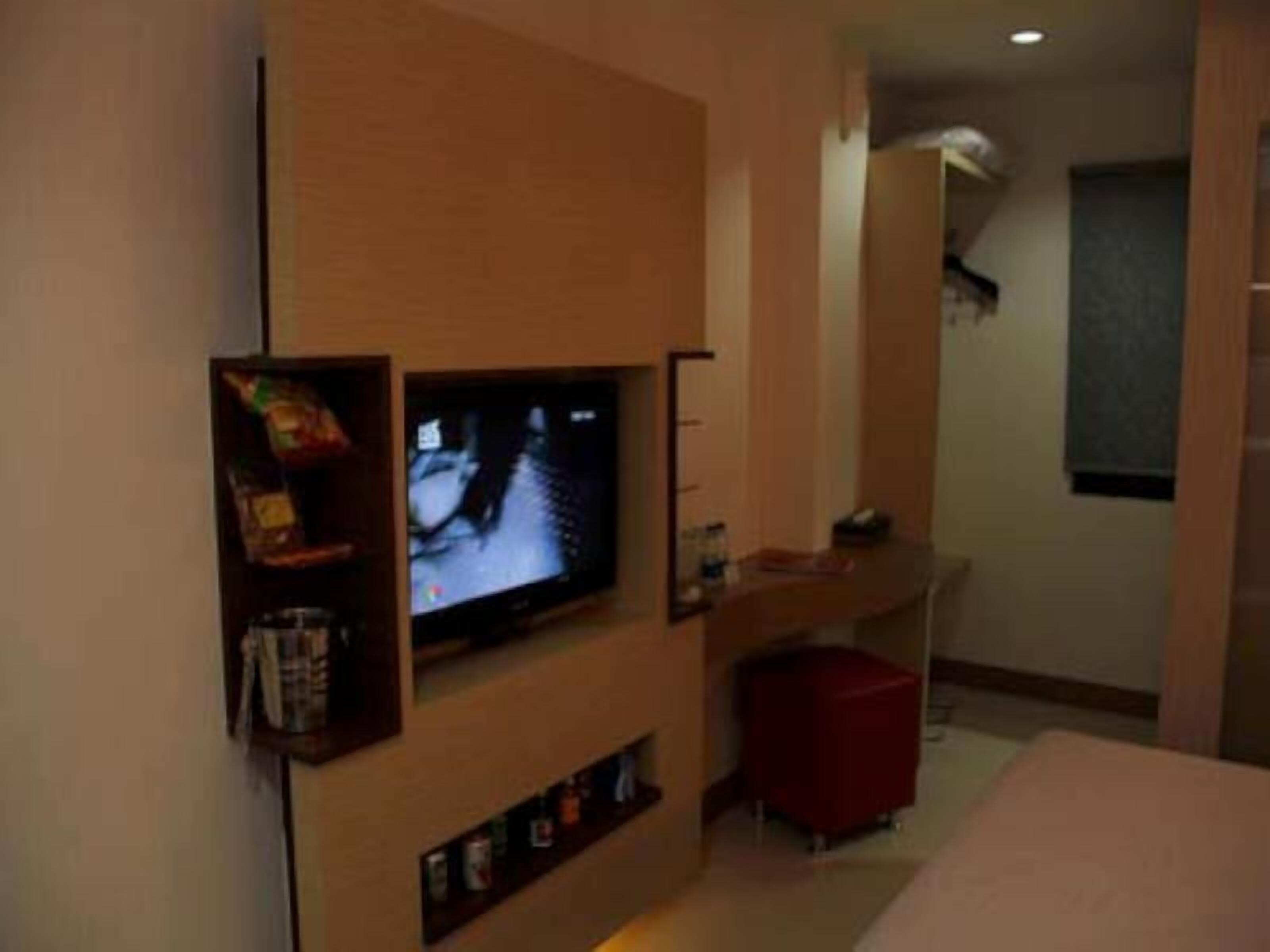 A modern hotel room with a flat-screen TV mounted on the wall, a small red ottoman, and a minimalist design.