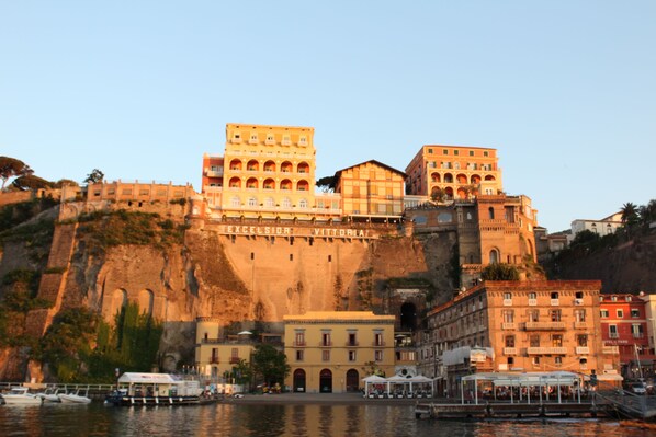 A coastal cityscape with historic buildings, including a prominent structure labeled "EXCELSIOR VITTORIA." - Surriento Suites (Sorrento)