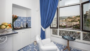 A room with a view of a coastal town, a white chair, a small table with a book, and a fruit bowl on a counter. - Surriento Suites (Sorrento)