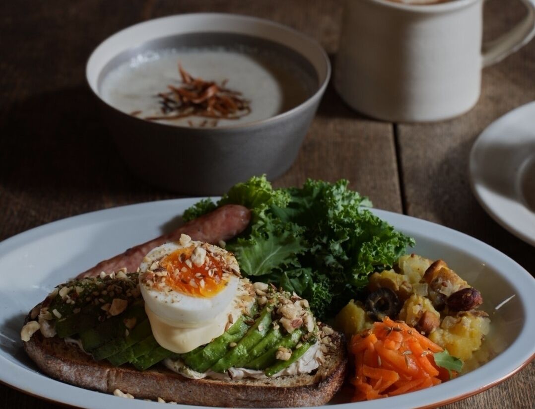A plate with toast topped with egg, greens, and nuts, alongside a bowl of soup and a cup of coffee on a wooden table.