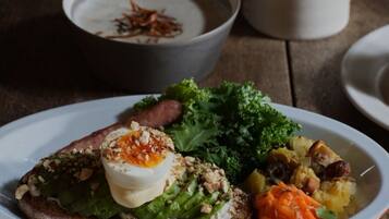 A plate with toast topped with egg, greens, and nuts, alongside a bowl of soup and a cup of coffee on a wooden table.