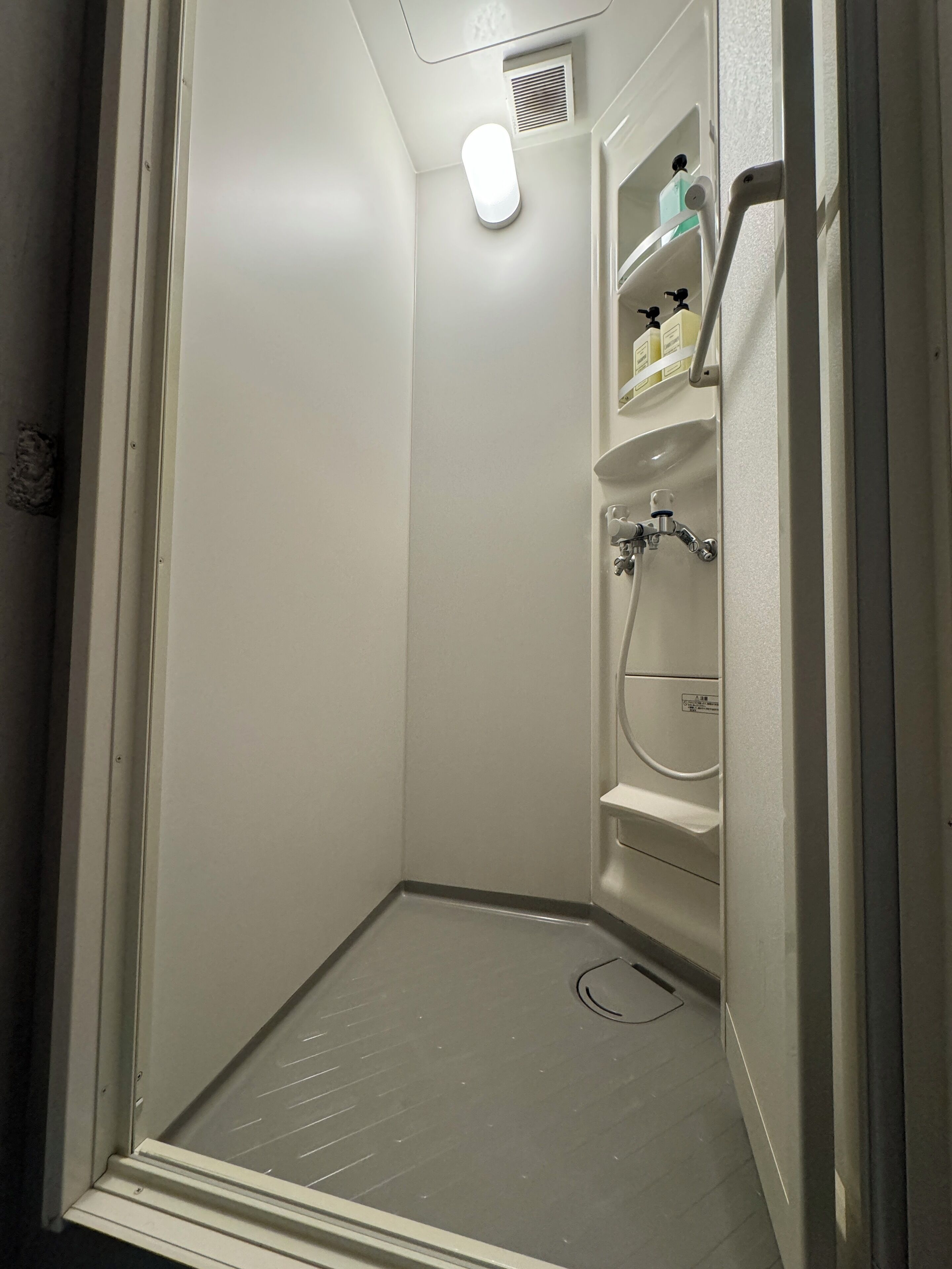 A corner shower with a white door, a shelf with toiletries, and a vent on the ceiling.