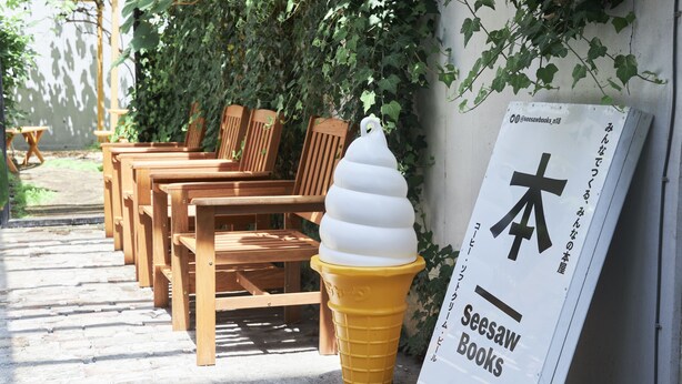A row of wooden chairs and a yellow ice cream cone sculpture outside a building with a sign for Seesaw Books. - Untapped Hostel (Sapporo)