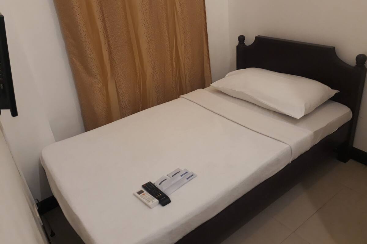 A single bed with a white mattress and a dark wooden headboard. A white pillow is placed on the bed.