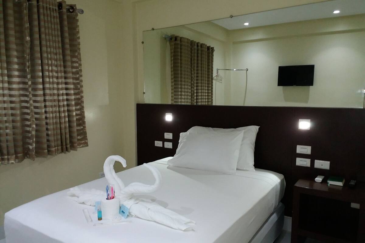 A hotel room with a neatly made bed, a bedside table with a phone and a book, a mirror above the bed, and a window with curtains.