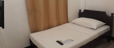 A single bed with a white mattress and a dark wooden headboard. A remote control and a magazine are placed on the bed.