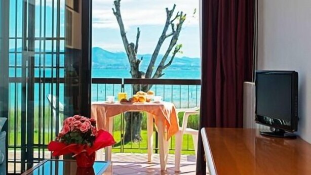 A room with a balcony, a table with a vase of flowers, and a TV on a wooden surface. - Virgilio Residence (Sirmione)