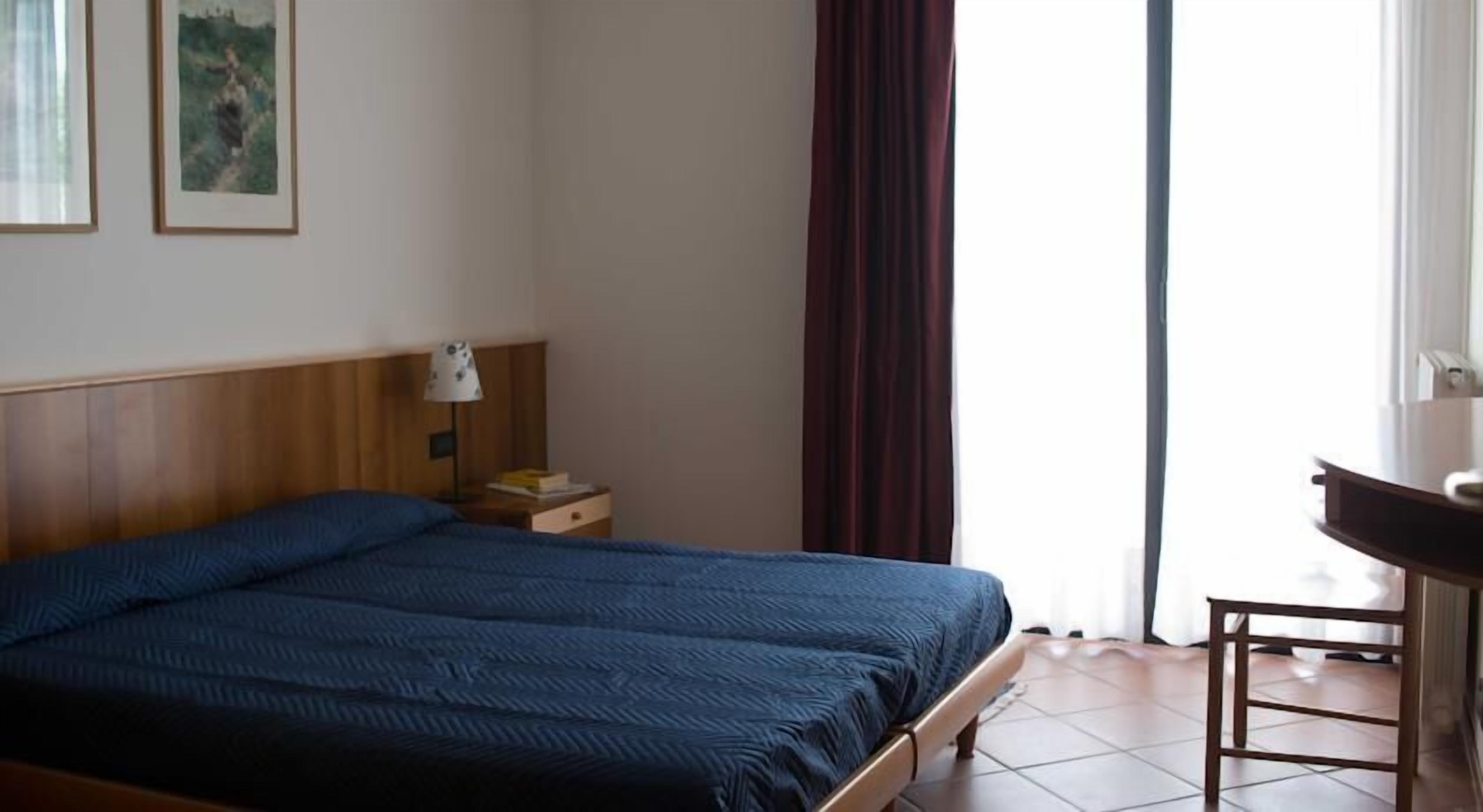 A bedroom with a bed, a nightstand, a lamp, a desk, and a window with curtains.