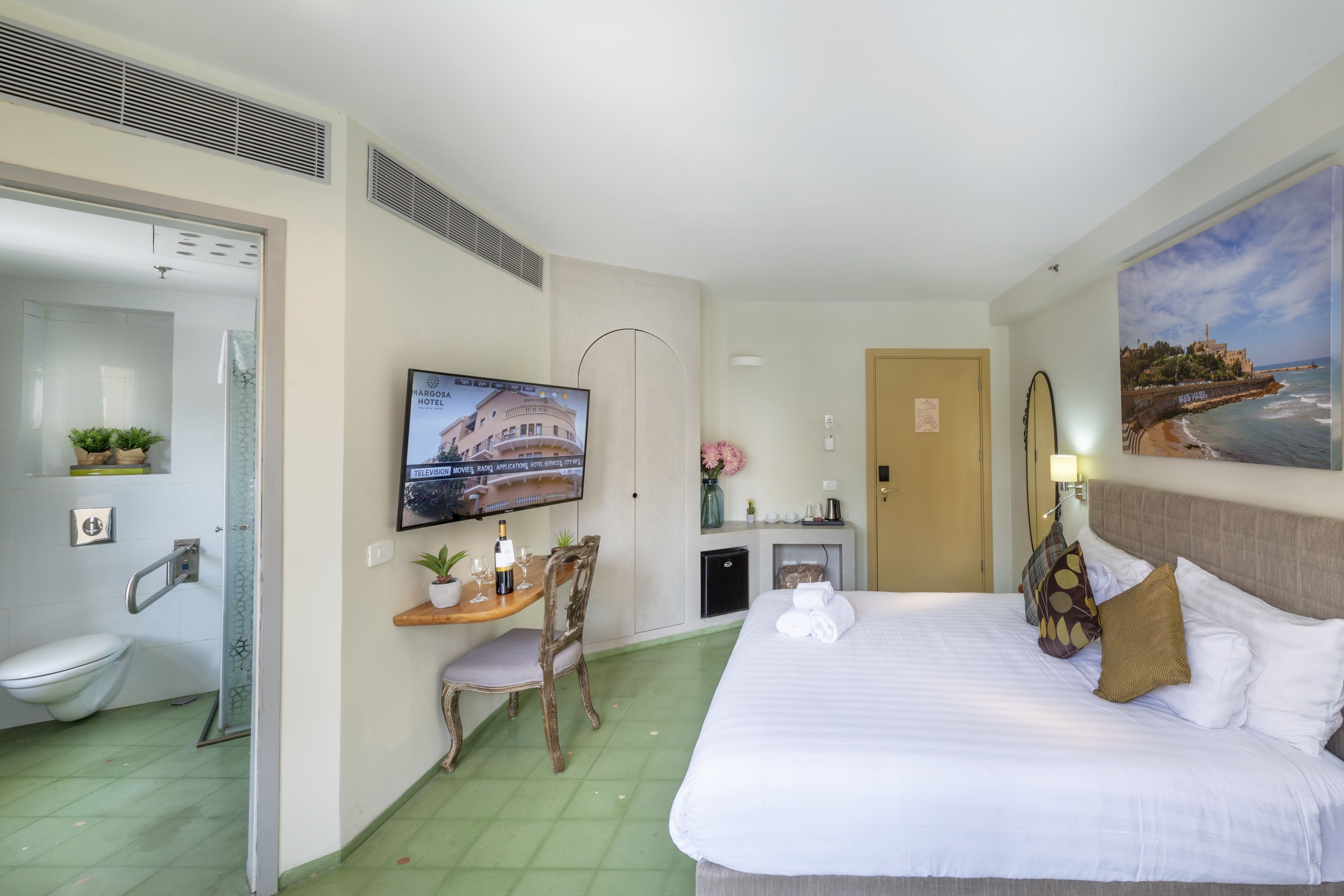 A hotel room with a bed, a desk with a chair, a television, a bathroom, and a painting on the wall.