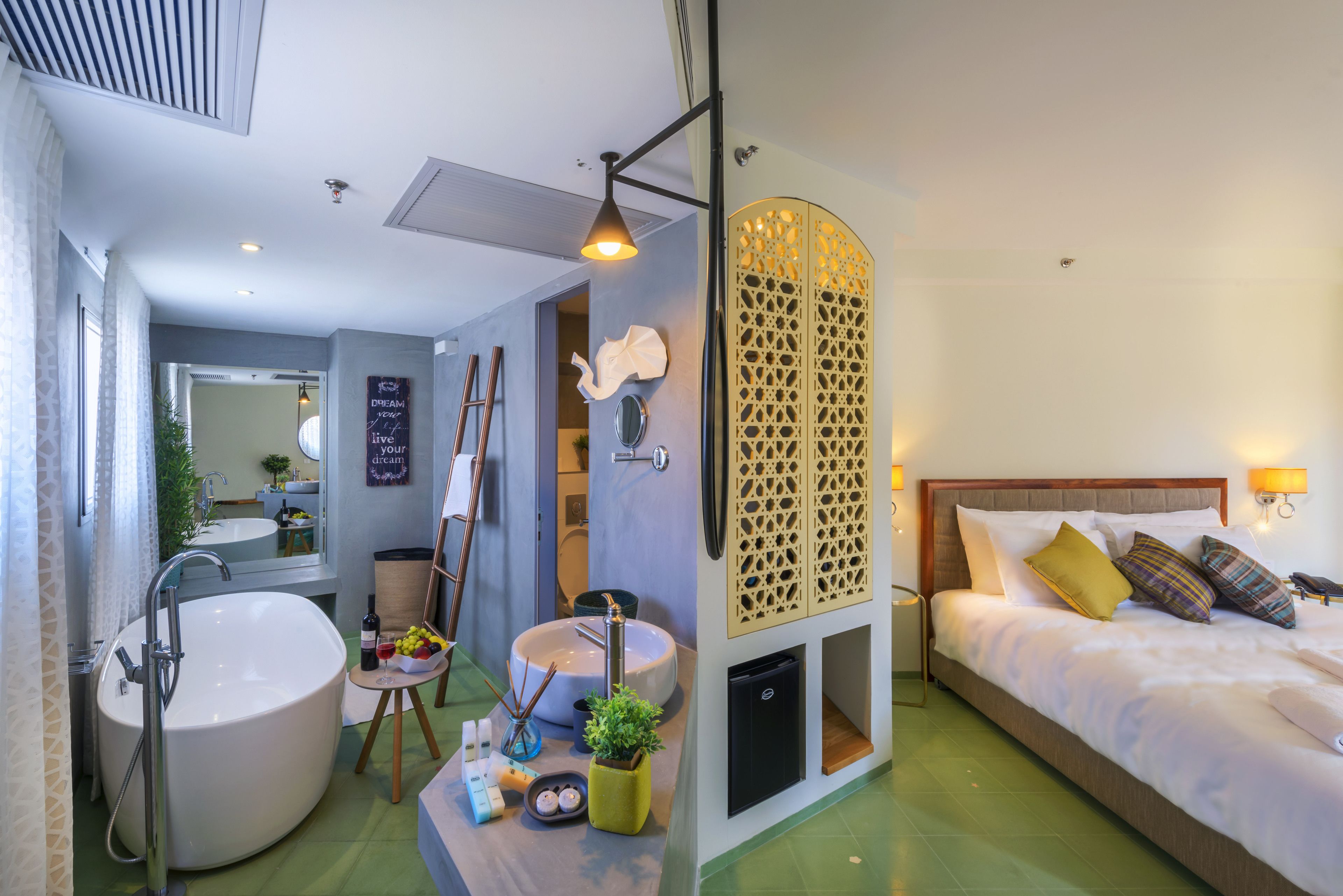 A modern hotel room with a large bathtub, a separate bathroom with a mirror and sink, a bed with multiple pillows, and a decorative wall panel.