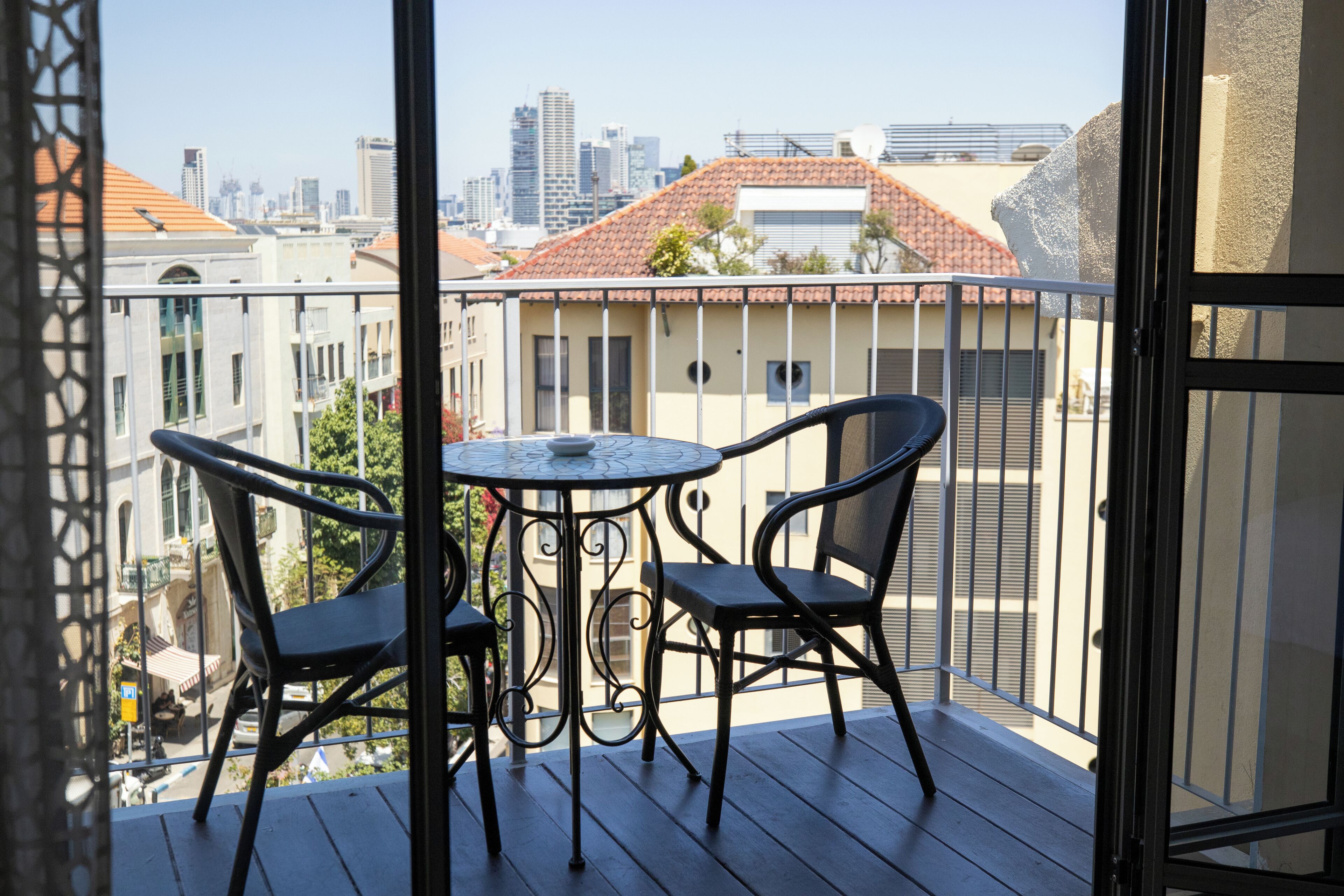 A balcony with a table and chairs, overlooking a cityscape with buildings and a clear sky.
