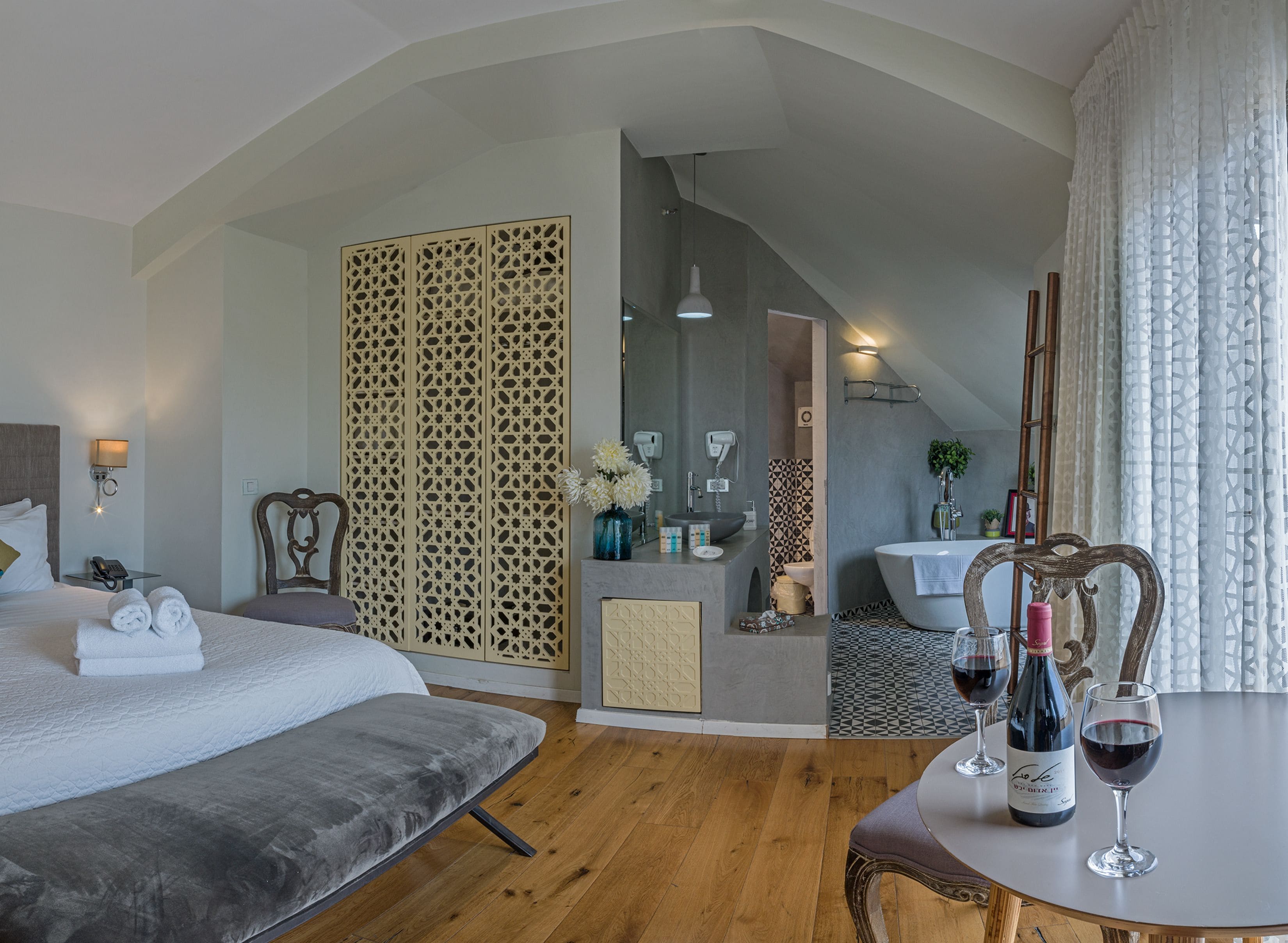 Habitación de hotel con cama, una mesita con copas de vino, baño con lavamanos y espejo, y un separador decorativo.