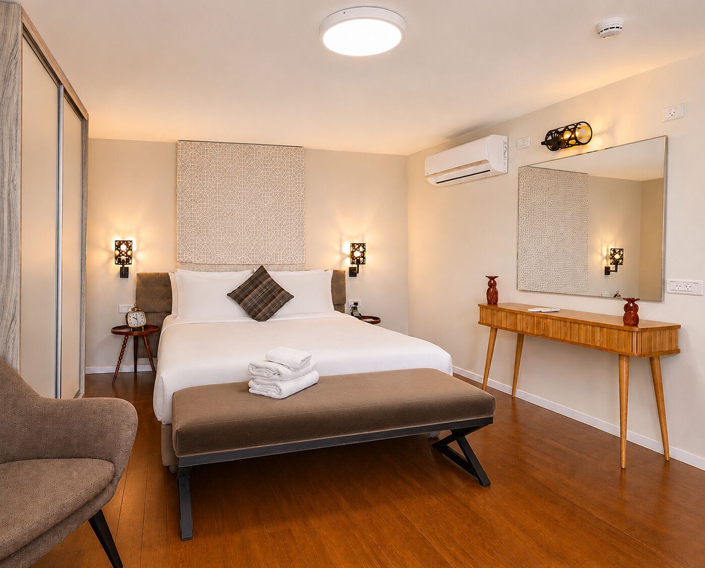 Una camera d'albergo con un letto, comodini, una sedia, una panca in legno, uno specchio e un condizionatore d'aria.