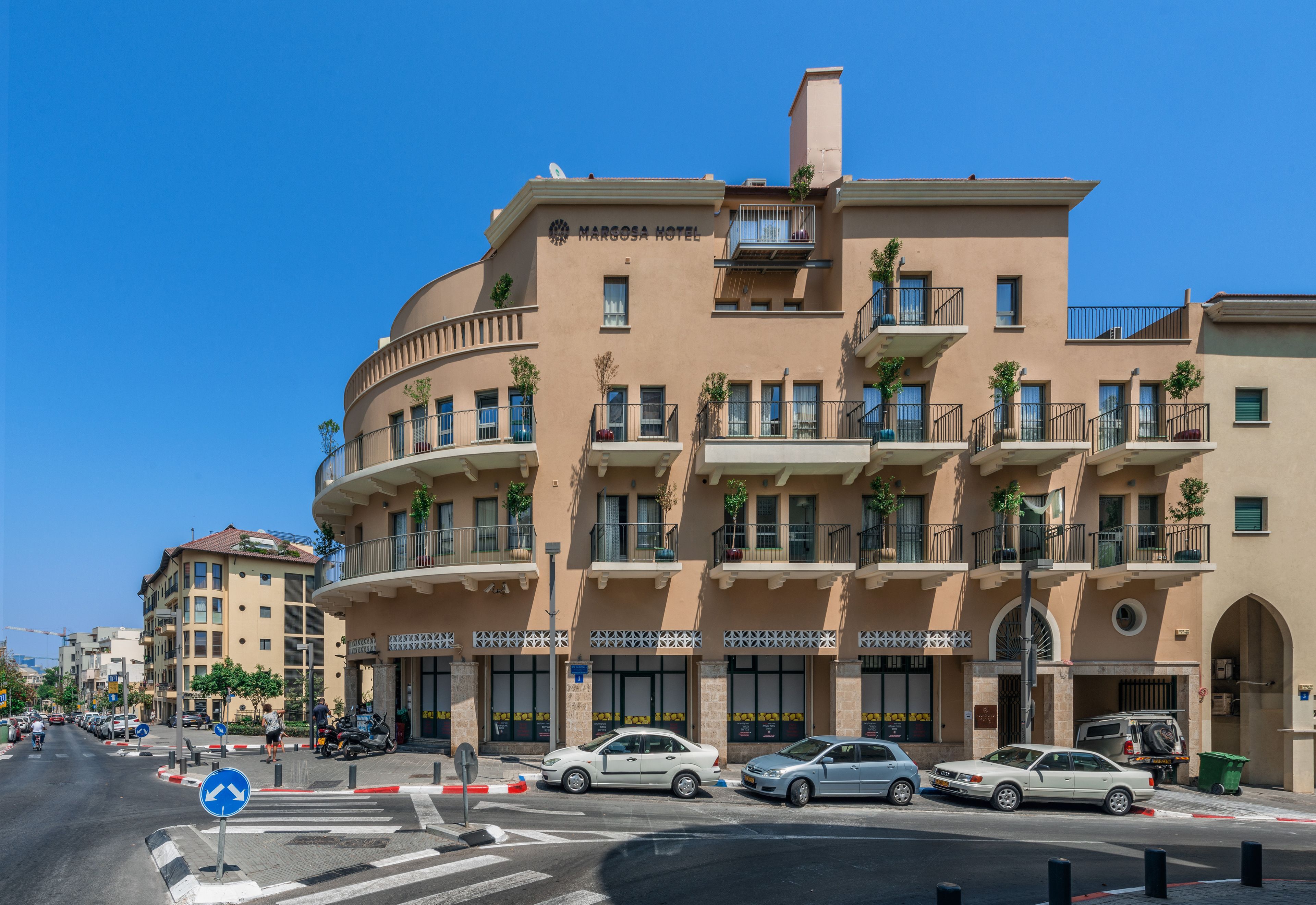 Un hotel de varios pisos con balcones, un cielo azul despejado y una calle con coches aparcados.
