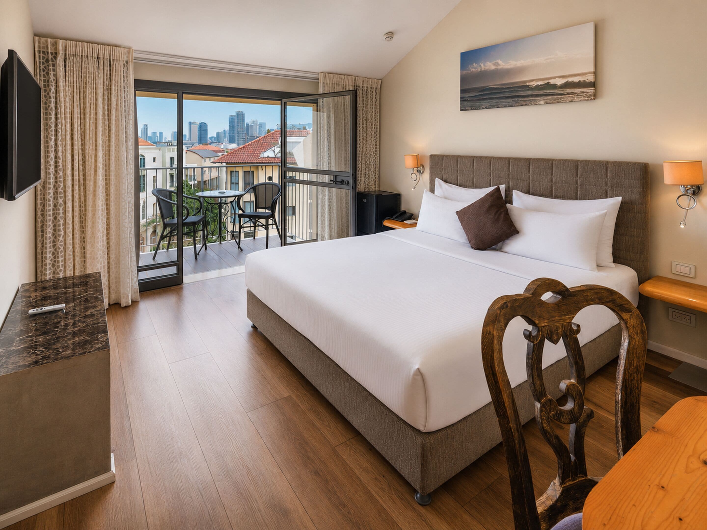 A hotel room with a large bed, a desk, a chair, a TV, a balcony with a view of the city, and a painting on the wall.