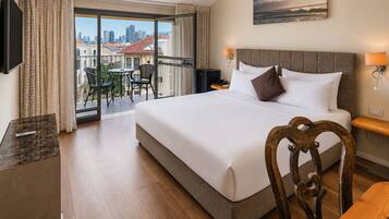 A hotel room with a large bed, a desk, a chair, a TV, a balcony with a view of the city, and a painting on the wall.