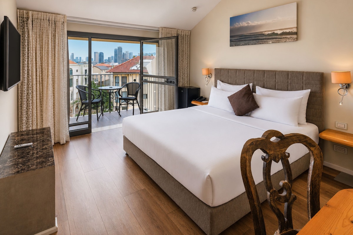 Habitación de hotel con una cama grande, un escritorio, una silla, televisión, balcón con vistas a la ciudad y un cuadro en la pared.