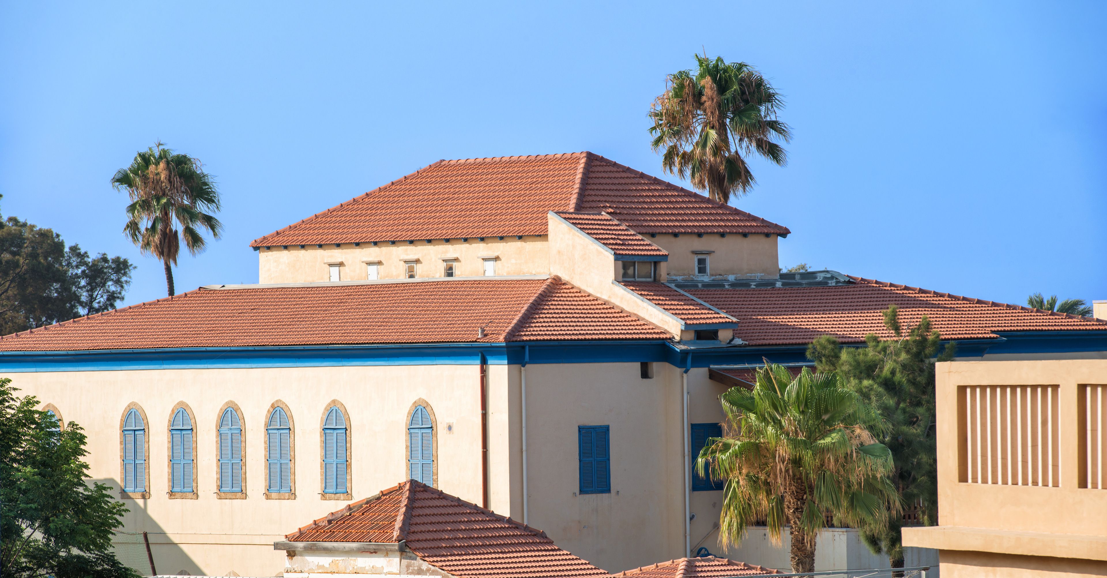 A building with a red-tiled roof, arched windows, and palm trees.
