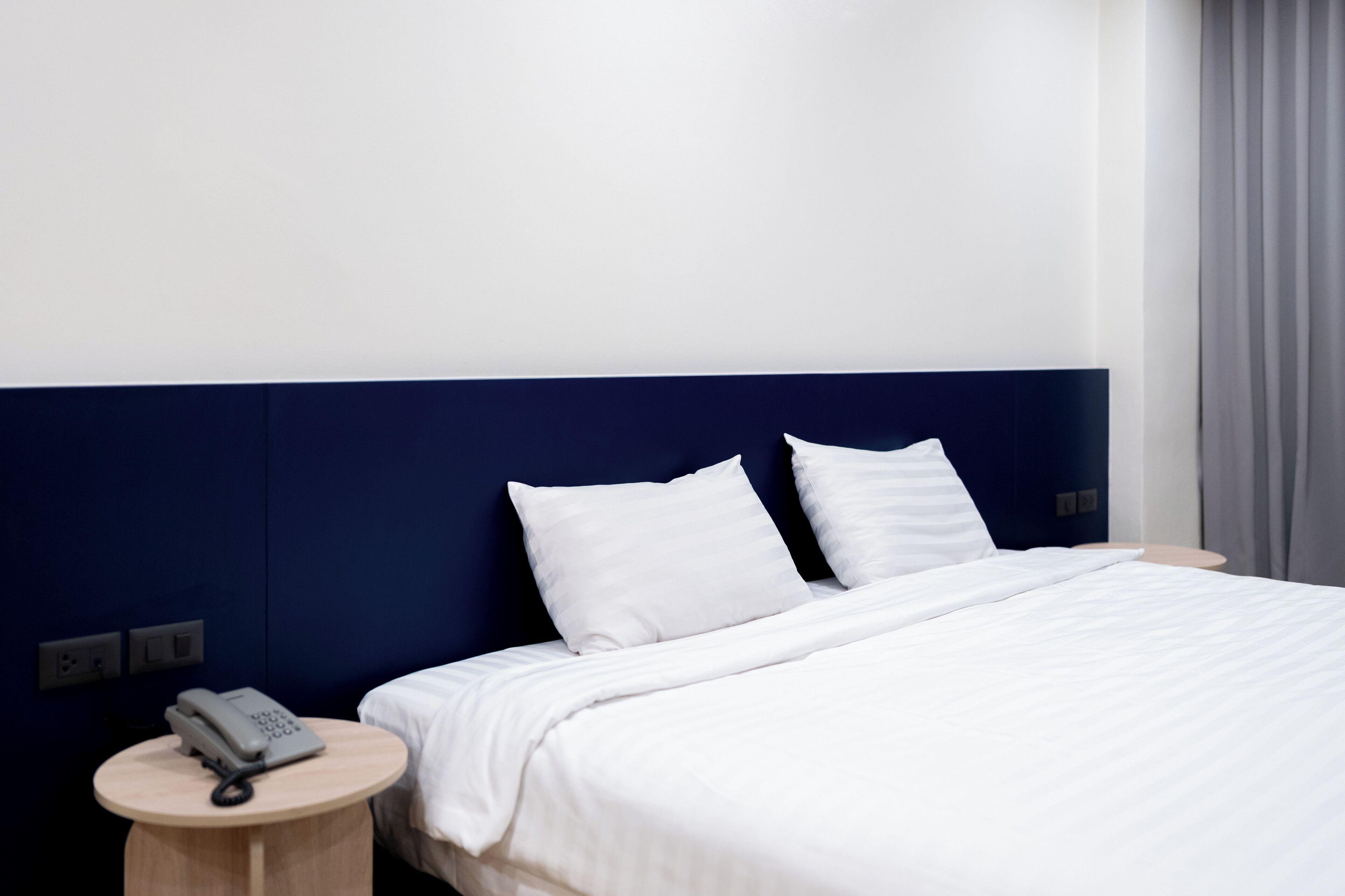Een hotelkamer met een bed, wit beddengoed, een rond houten nachtkastje met een telefoon en een donkerblauwe hoofdbord.